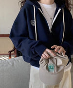 ネイビーのハーフジップパーカーを着用した女性が、ベージュのキャップを持ち、ソファの前に立っている。インナーは白Tシャツ、ボトムスは生成り色のワイドパンツ。