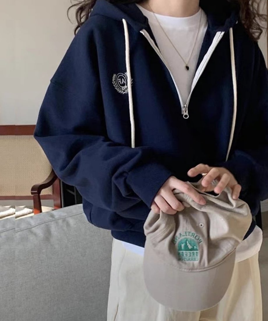 ネイビーのハーフジップパーカーを着用した女性が、ベージュのキャップを持ち、ソファの前に立っている。インナーは白Tシャツ、ボトムスは生成り色のワイドパンツ。