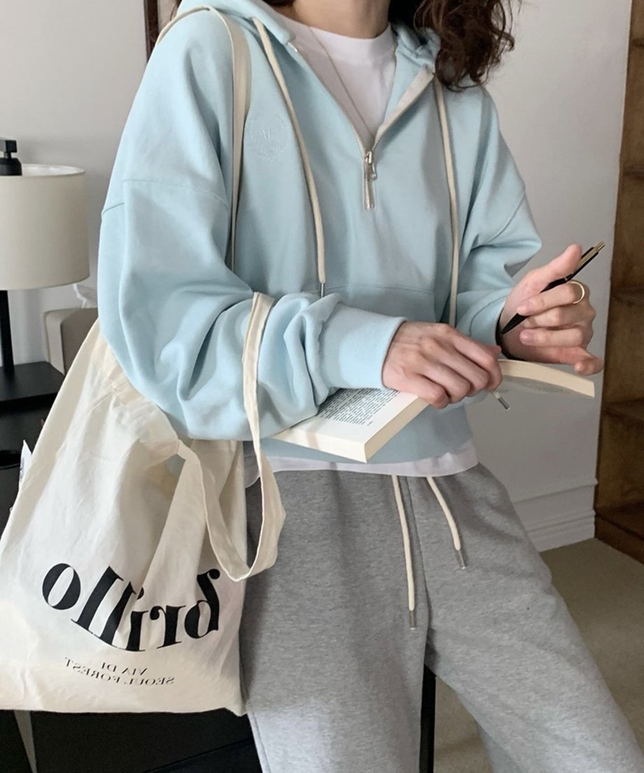 ミントグリーンのハーフジップパーカーを着用した女性の上半身ショット。白いTシャツをインナーに、グレーのスウェットパンツと白いロゴ入りトートバッグを合わせ、本を持っている。