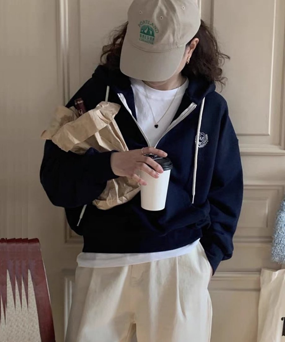 ネイビーのハーフジップパーカーを着て、茶色の紙袋に入ったワインボトルを抱えている女性のクローズアップ。白いTシャツをインナーに着ており、首元に黒のペンダントが見える。
