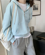ミントグリーンのハーフジップパーカーを着用した女性が、グレーのスウェットパンツと白いロゴ入りトートバッグを合わせて室内に立っている。