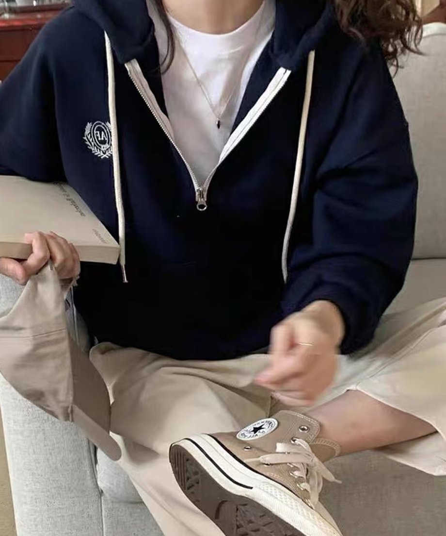 ネイビーのハーフジップパーカーを着用した女性が椅子に座っている。インナーは白Tシャツ、ボトムスは生成り色のワイドパンツで、手にキャップを持ってベージュのハイカットスニーカーを履いている。