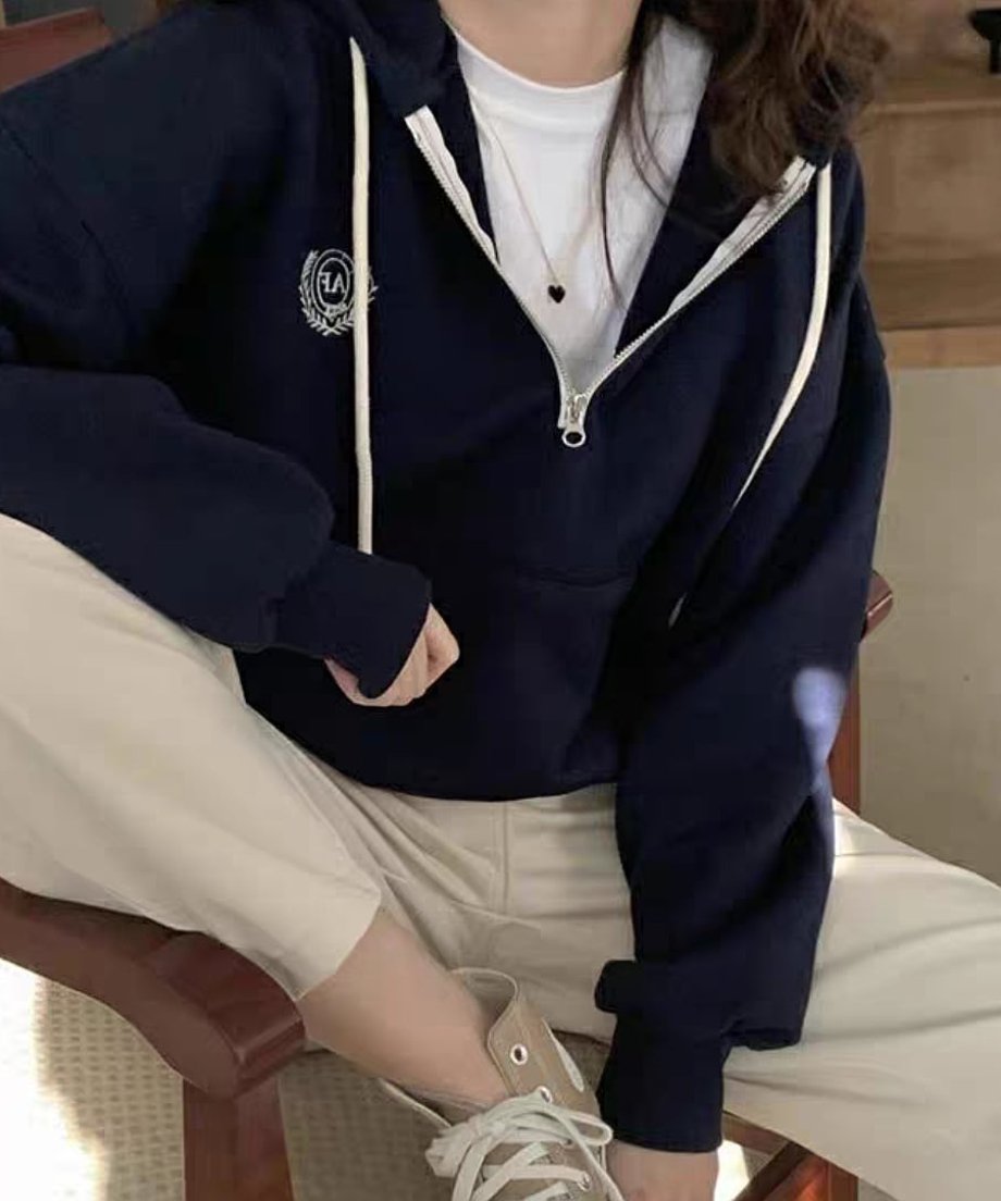 ネイビーのハーフジップパーカーを着用した女性が椅子に座っている。インナーは白Tシャツ、ボトムスは生成り色のワイドパンツで、ベージュのハイカットスニーカーを履いている。
