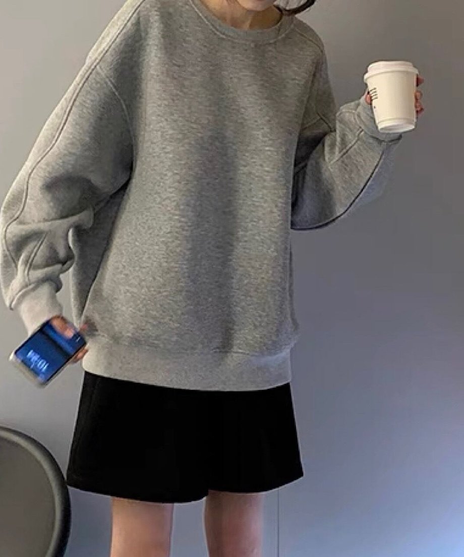 グレーのオーバーサイズスウェットを着用し、黒のショートパンツを合わせた女性。片手にスマートフォン、もう一方の手に紙コップを持っている。