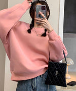 ピンクのスウェットを着用した女性のセルフィー。スマートフォンで撮影しており、眼鏡を頭に乗せ、複数のリングとネックレスを着用している。