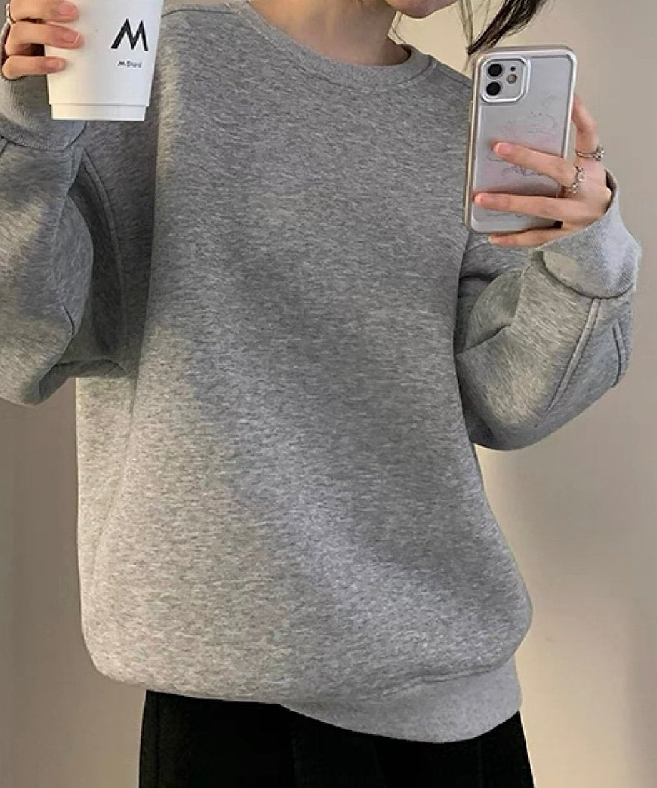 グレーのオーバーサイズスウェットを着用した女性の全身。黒のボトムスを着用し、シンプルなカジュアルスタイルです。手にはカップとスマホを持っている。