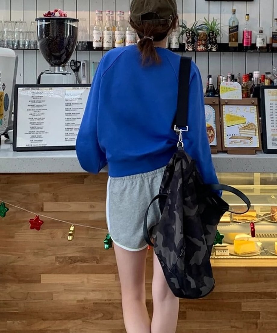 ブルーのVネックプルオーバーとグレーのショートパンツを着用し、カフェのカウンターに立っている女性の後ろ姿。