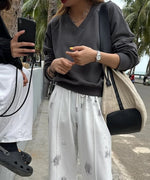 グレーのVネックスウェットと白いタイダイ柄のイージーパンツを着用し、屋外でスマートフォンを見ている女性。