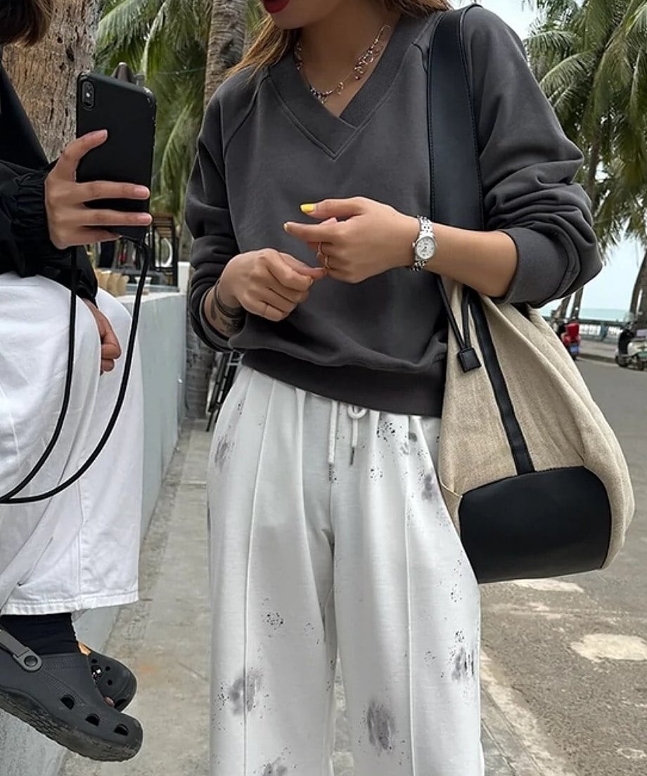 グレーのVネックスウェットと白いタイダイ柄のイージーパンツを着用し、屋外でスマートフォンを見ている女性。