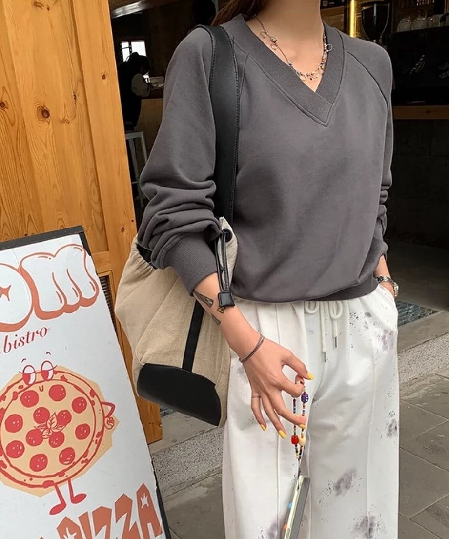グレーのVネックスウェットを着た女性が、カラフルなビーズのブレスレットを持っている。カフェの入口に立っている。