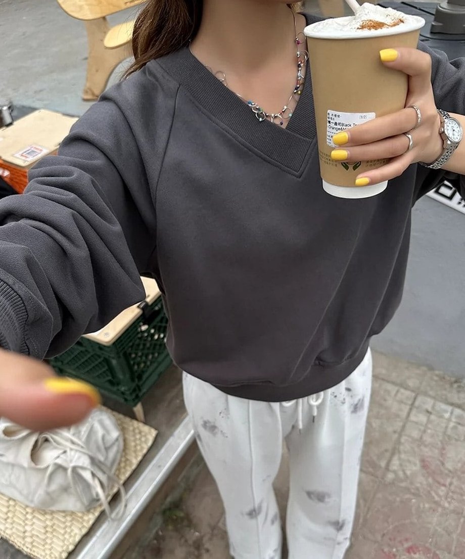 Vネックのチャコールグレースウェットシャツを着た女性が屋外でコーヒーカップを持っている写真。