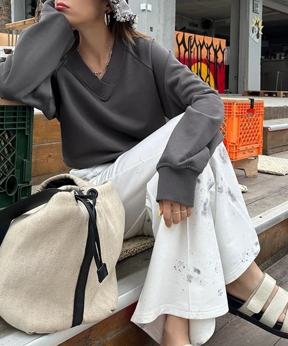グレーのVネックスウェットと白いタイダイ柄パンツを着用し、屋外の木製ベンチに座っている女性。