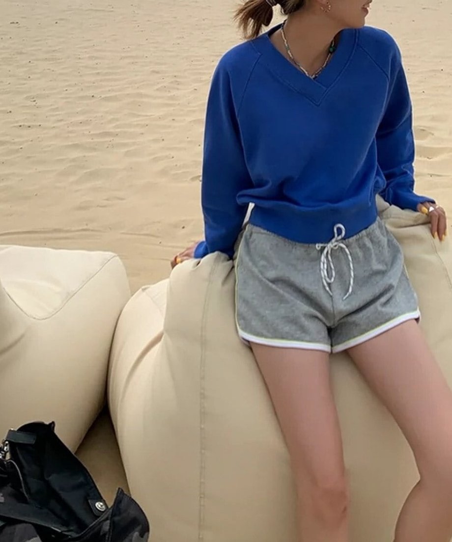 ブルーのオーバーサイズスウェットとグレーのショートパンツを着用し、ビーチサイドのソファに座る女性。