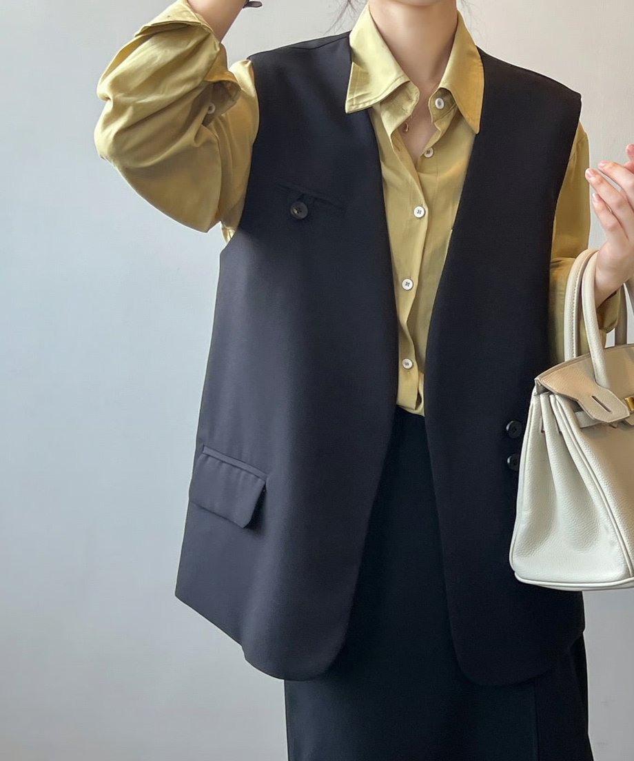 黒のVネックベストを淡い黄色の長袖シャツと黒のスカートに合わせた、きれいめな着こなしの女性の上半身。片手をあげて白いハンドバッグを持っている。