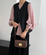 黒のワンボタンクロスベストをスモーキーピンクのブラウスとブラックのスカートに合わせた女性の上半身。茶色のハンドバッグを持っている。