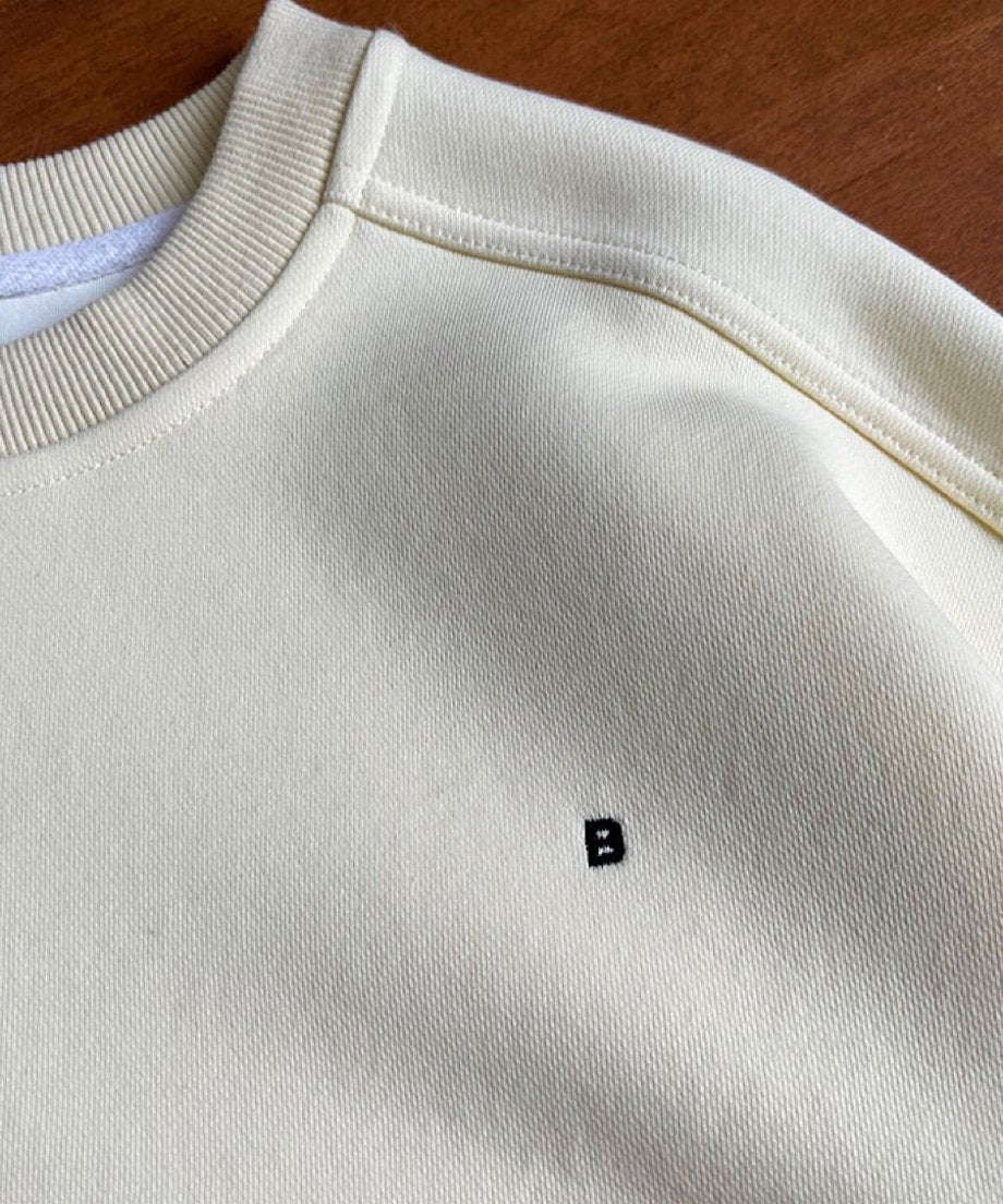 クリームのオーバーサイズスウェットの襟から肩にかけての接写。胸元のBのマークがわかる。