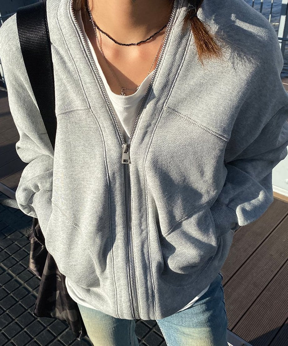 屋外の橋の上で、明るいグレーのオーバーサイズVネックジップアップスウェットカーディガンを着用した女性の正面クローズアップ。白いTシャツをインナーに、ジップを半開けにし、両手をポケットに入れている。