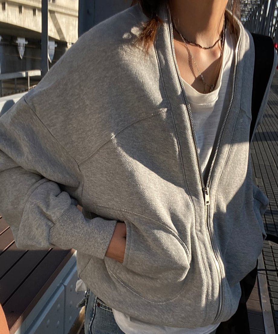 屋外の橋の上で、明るいグレーのオーバーサイズVネックジップアップスウェットカーディガンを着用した女性の正面クローズアップ。白いTシャツをインナーに、ジップを半開けにし、両手をポケットに入れている。
