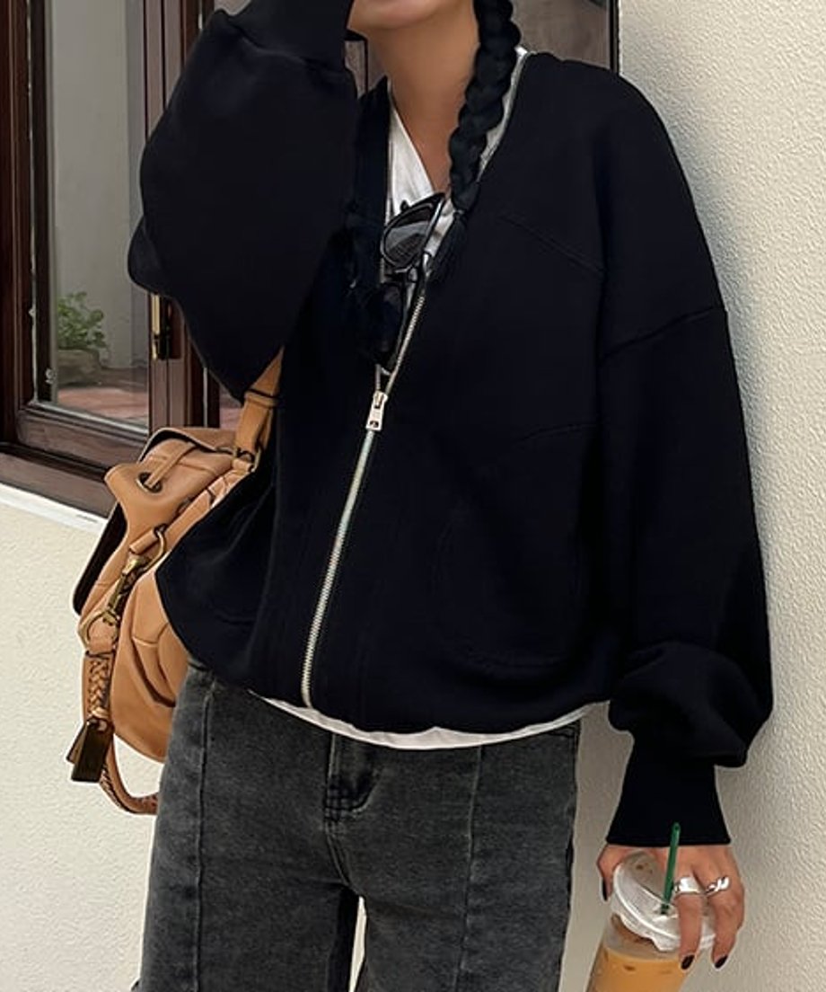 外壁の前で、黒のオーバーサイズVネックジップアップスウェットカーディガンを着用した女性。ボトムスは濃いグレーのデニムパンツ、インナーは白Tシャツ、キャメル色のバックパックを合わせている。