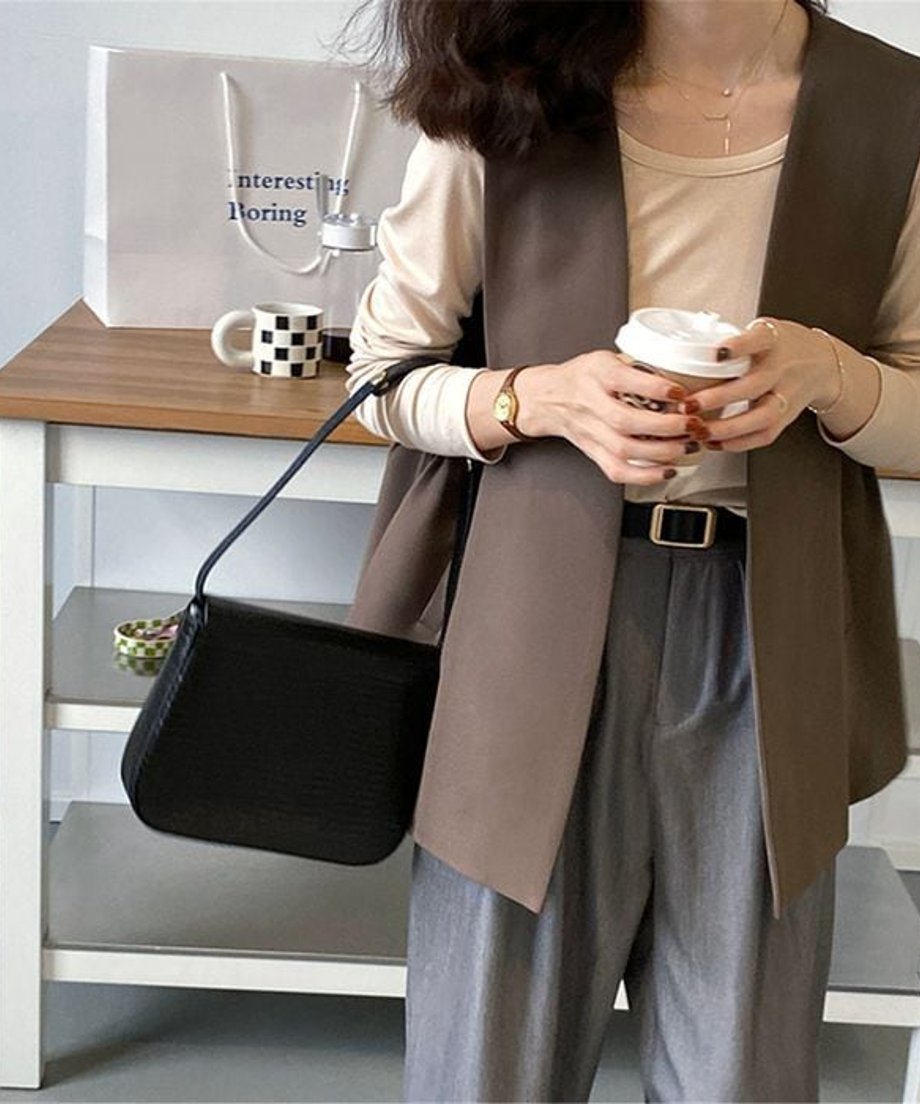 ブラウンのロングベストを着用した女性の正面。薄いベージュの長袖Tシャツとグレーのスラックスに黒のベルトを合わせ、縦のラインを強調したすっきりとした着こなし。