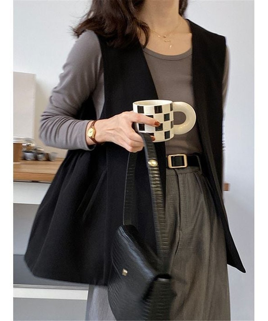 ブラックのロングベストを着用した女性の正面。ダークベージュの長袖Tシャツとグレーのスラックスを合わせ、黒のベルトと黒のショルダーバッグをコーディネートしている。手に黒と白のチェック柄のカップを持っている。