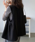 黒のロングベストを着用した女性の正面。グレーの長袖Tシャツとグレーのスラックスに黒のベルトを合わせ、チェック柄のマグカップを持っている。
