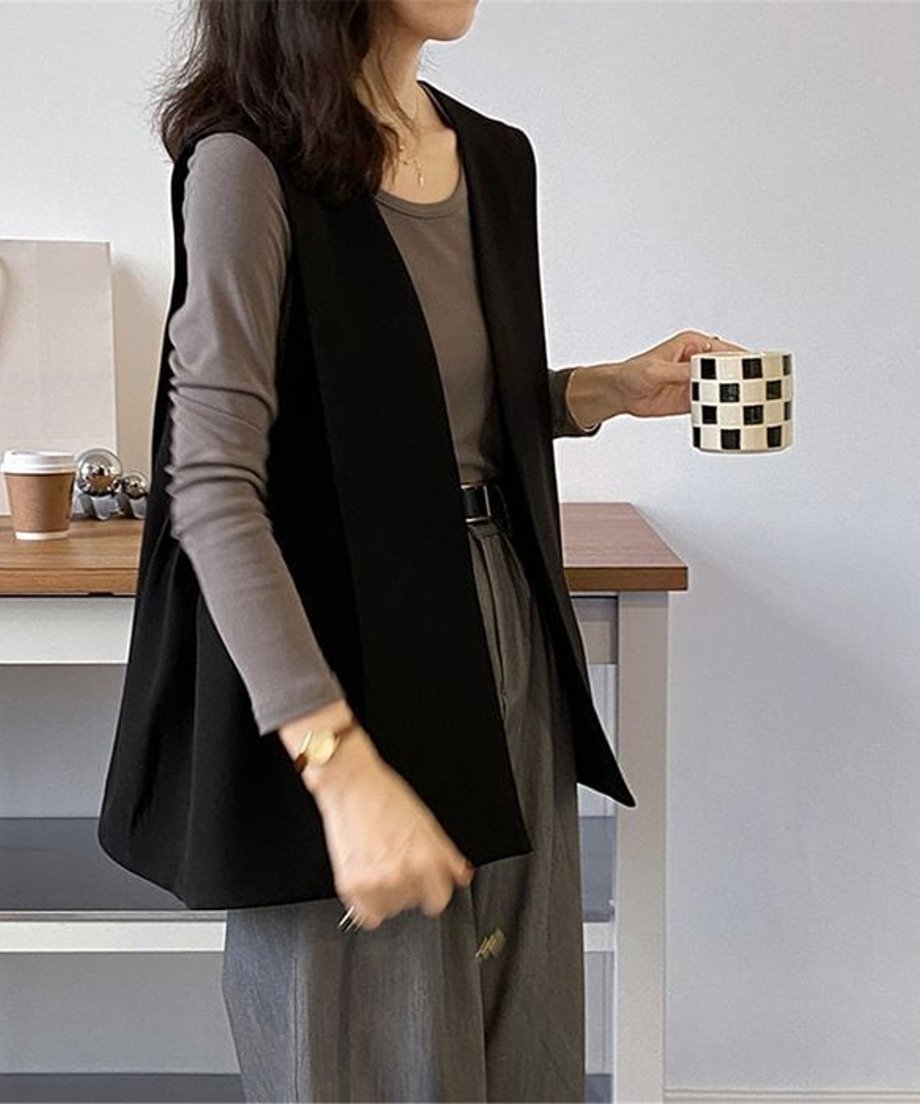 ブラックのロングベストを着用した女性の正面。チャコールの長袖Tシャツとグレーのスラックスを合わせ、黒のベルトをコーディネートしている。手に黒と白のチェック柄のカップを持っている。