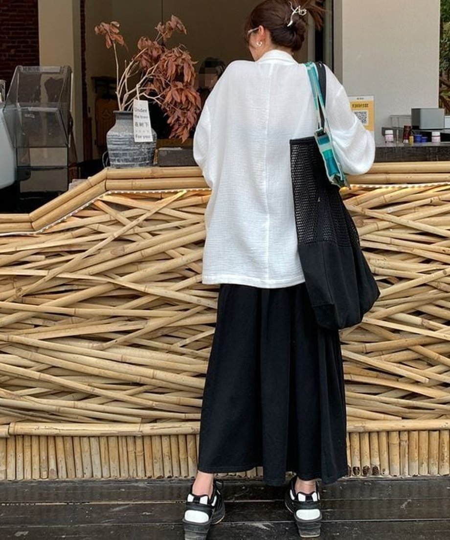 白のルーズジャケットを後ろ姿で着用した女性。黒のロングスカートと厚底サンダルを合わせ、メッシュバッグを肩にかけている。