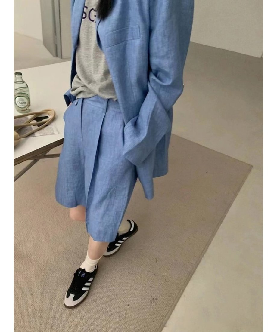 ブルーのリネン混ジャケットとバミューダパンツのセットアップを着用した女性。インナーにはグレーのロゴTシャツ、足元には黒のスニーカーを合わせたカジュアルなコーディネート。
