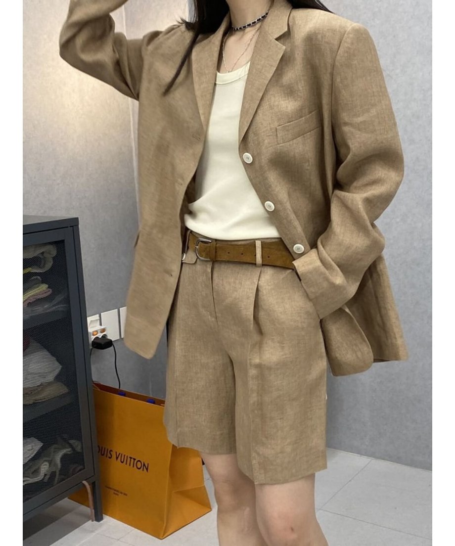 カーキのリネン混セットアップを着用した女性。白いタンクトップとブラウンのレザーベルトを合わせた室内ショット。