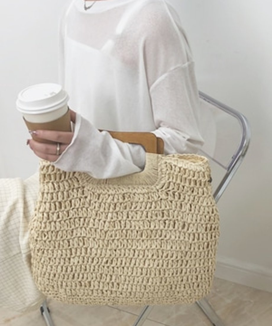 コーヒーを持ちながらアイボリーのストローバッグを腕に掛けて座る女性。