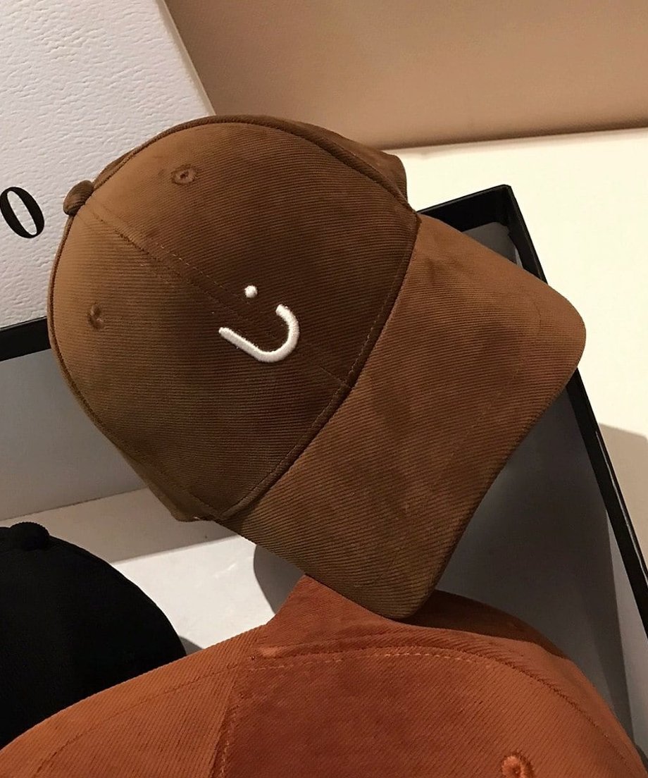 ダークブラウンのキャップを斜めから撮影。コーデュロイの細かい質感が際立ち、フロントのUの刺繍が強調されています。手前にはキャメル、ブラックのキャップが見える。