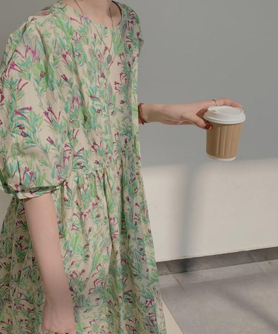 グリーンのボタニカル柄ワンピース。ふんわりとしたパフスリーブとラウンドネックのデザイン。手にドリンクを持った画像。