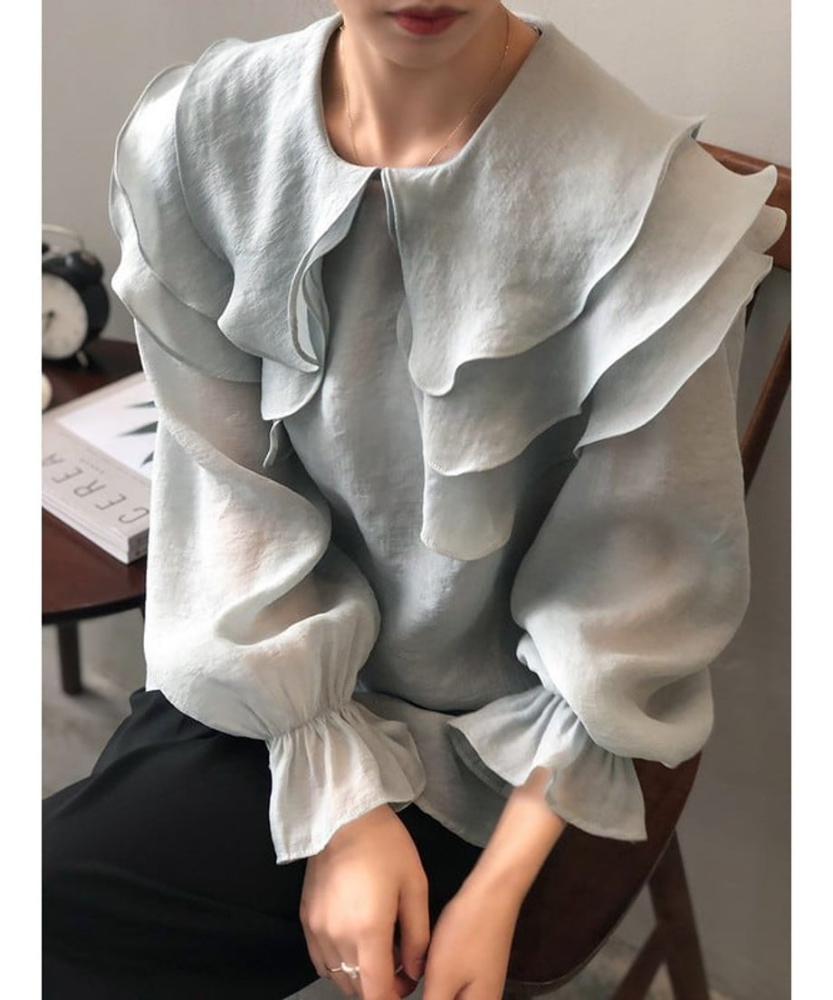 ブルーのティアードフリルブラウスを着用し、椅子に座る女性の正面アップ写真。