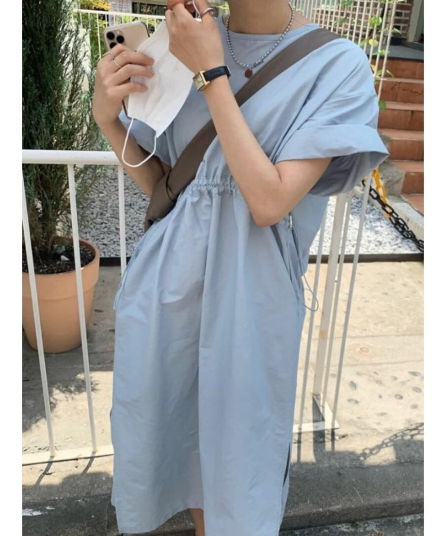 ブルーのワンピースを着用し、マスクを手に持つ女性。首元のネックレスと腕時計がアクセント。