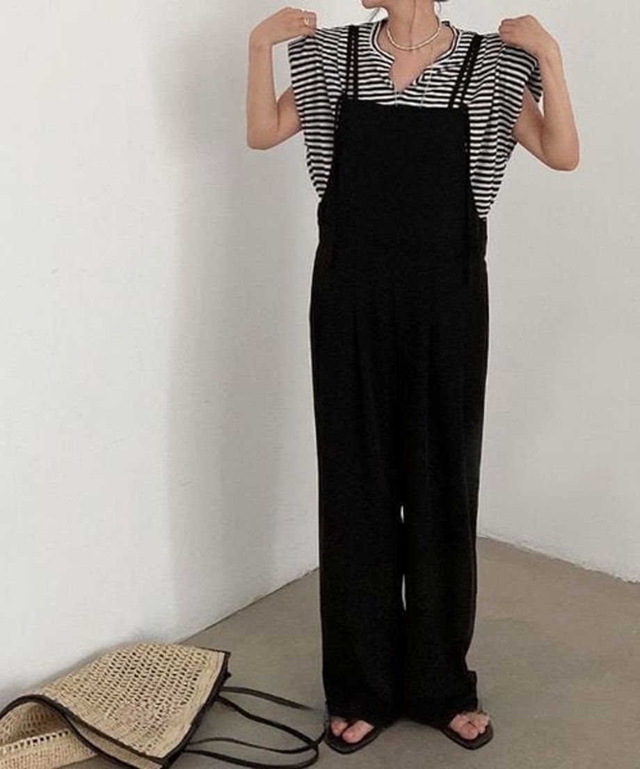 Vネックのボーダーシャツにダブルストラップのサロペットを合わせた女性が両肩を手で触れた写真。足元にはメッシュの大きなバッグが無造作に置かれている。