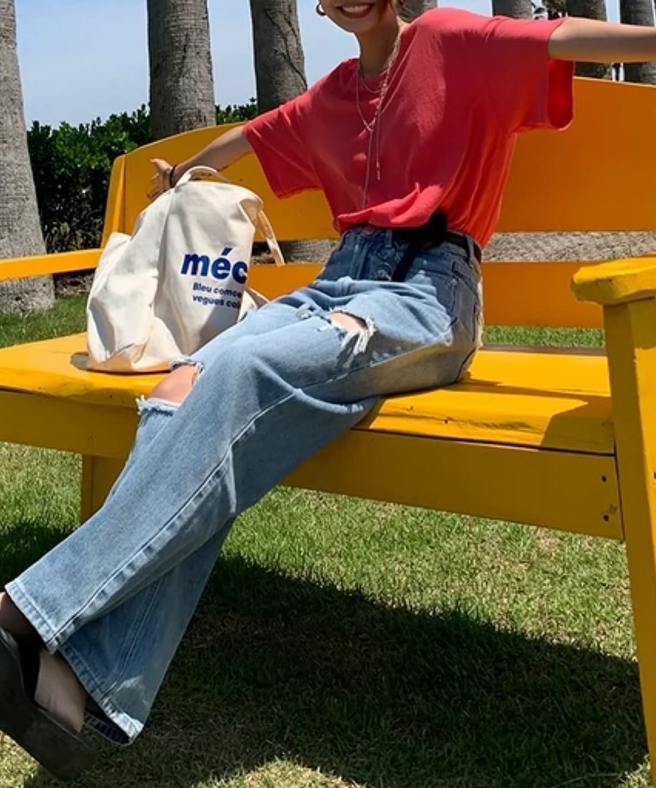 黄色いベンチに座り、ピンクレッドのTシャツとライトブルーのデニムを着用したリラックスした様子。
