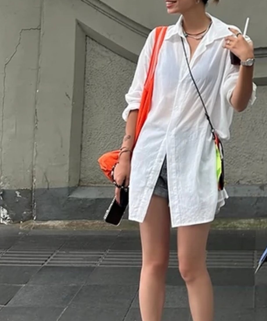 白の透け感があるシャツにショートパンツを合わせ、都会の街角に佇む女性のスタイリッシュな姿。