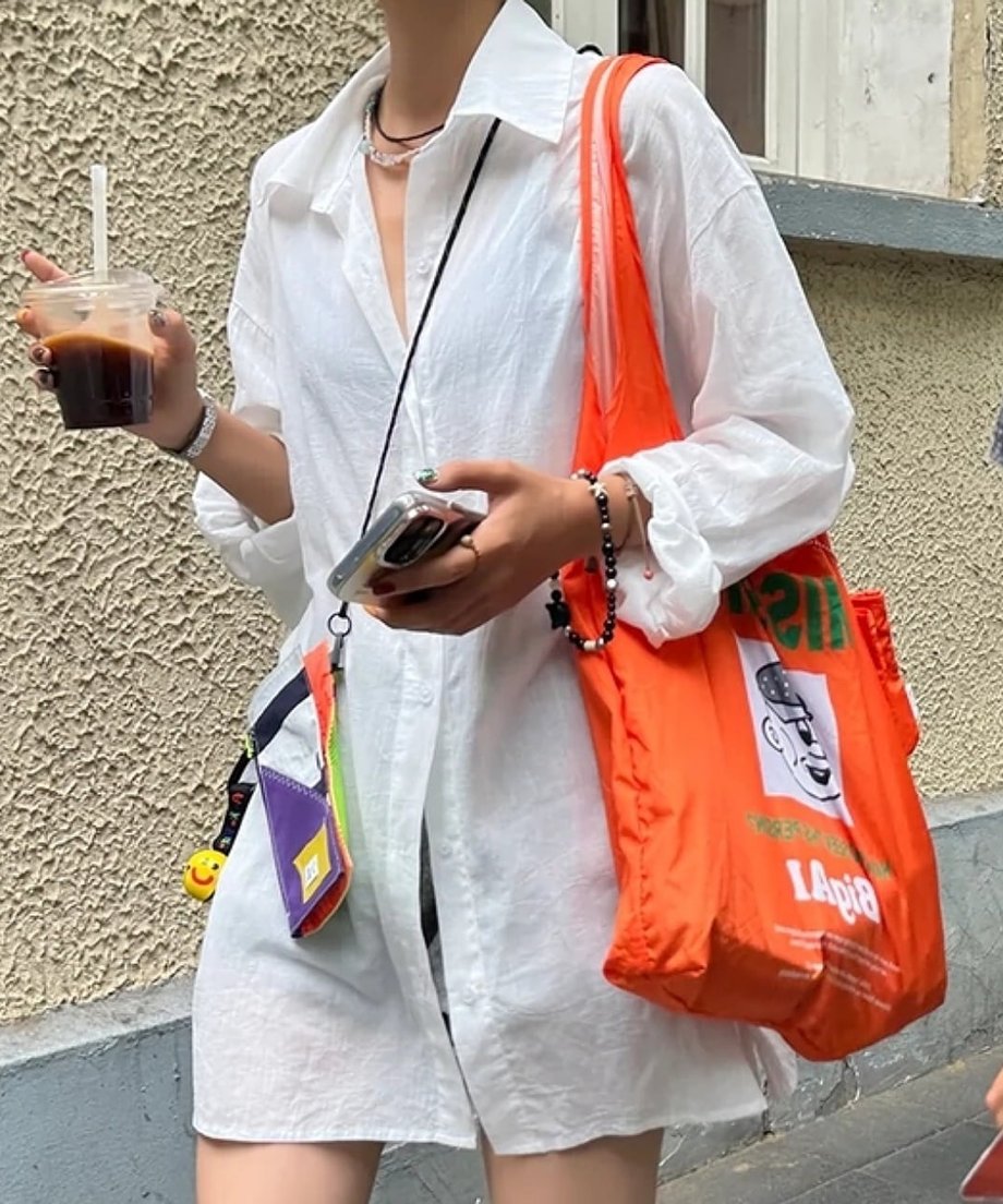 白のシャツを羽織り、手にコーヒーとスマートフォンを持って歩く街中でのカジュアルなスタイリング。