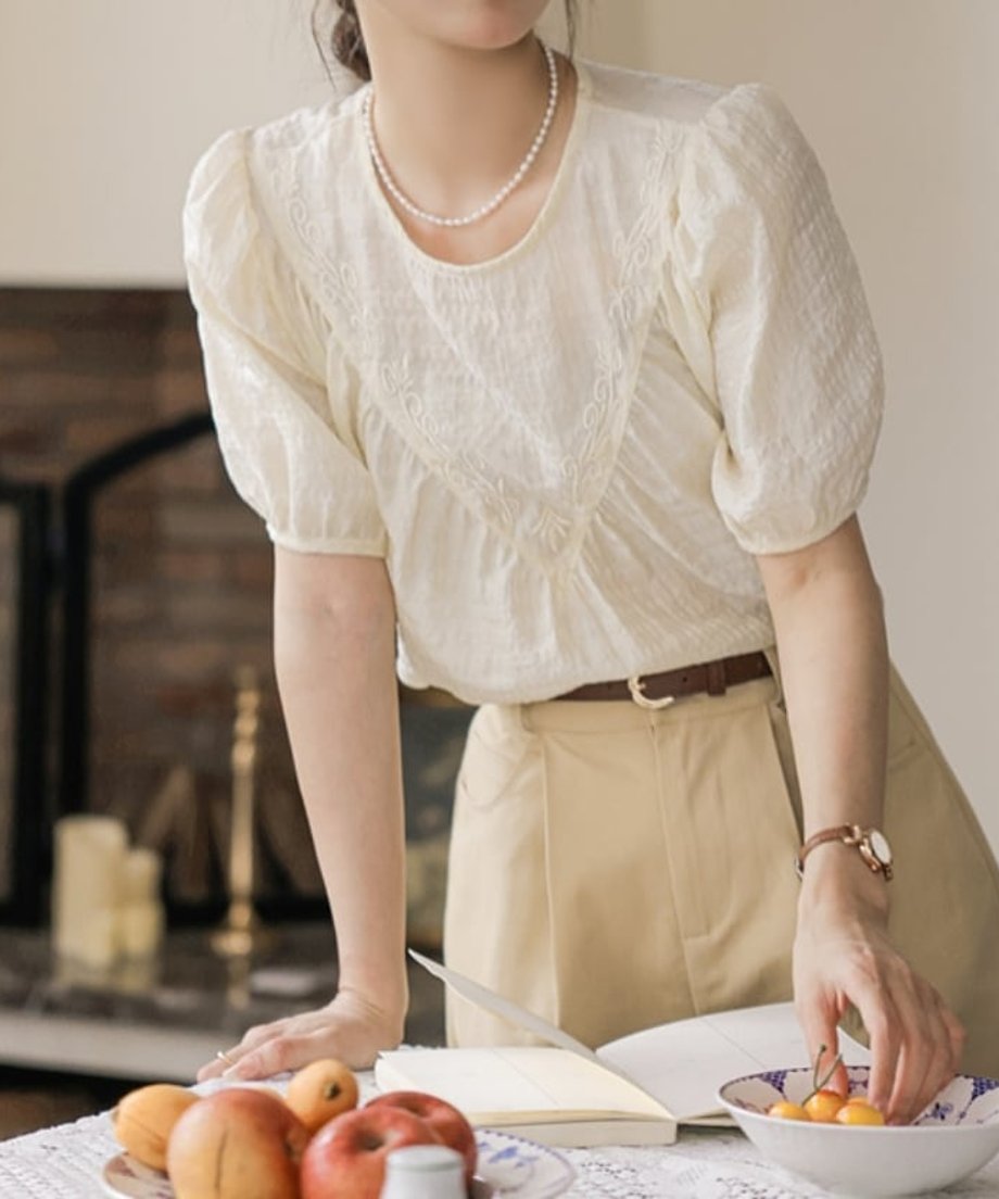アプリコットの刺繍ブラウスをベージュのボトムスに合わせ、朝食のシーンを楽しむ女性の様子
