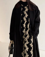 黒いロングコートの下に花柄のジャンパースカートと黒いタートルネックを着用した女性の上半身のクローズアップ画像。