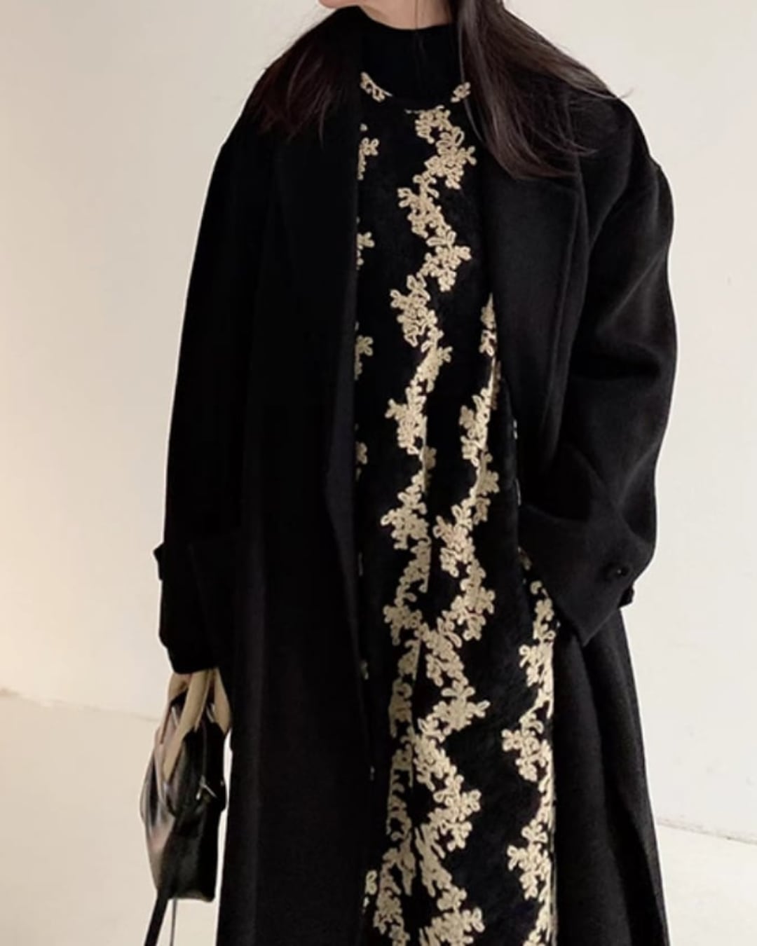 黒いロングコートの下に花柄のジャンパースカートと黒いタートルネックを着用した女性の上半身のクローズアップ画像。