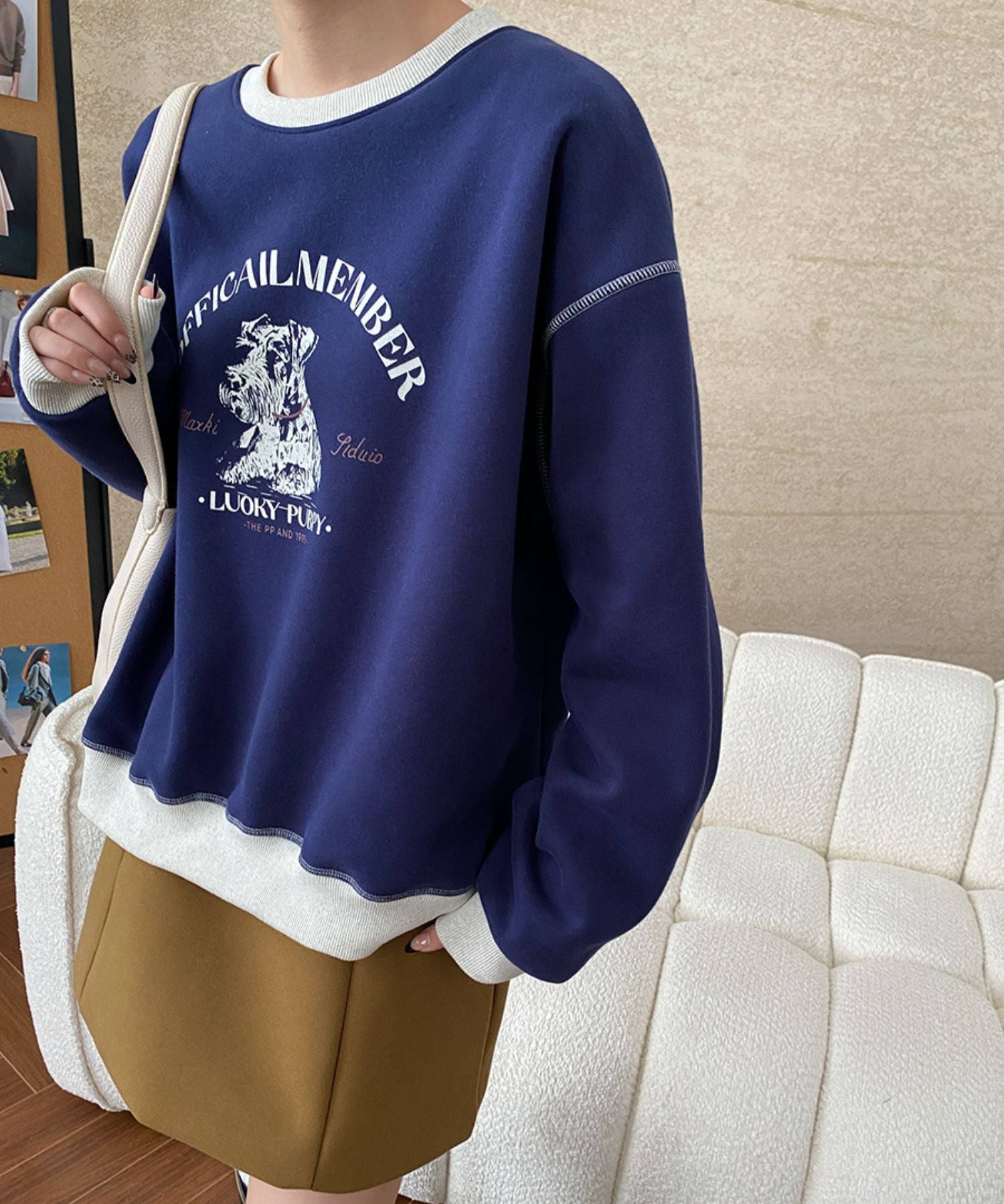 ブルーのボディに白のグラフィックがプリントされ、襟元、袖口、裾が白で縁取られたスウェットシャツを着用した人物。ブラウンのミニスカートと合わせてソファーの前に立っている。