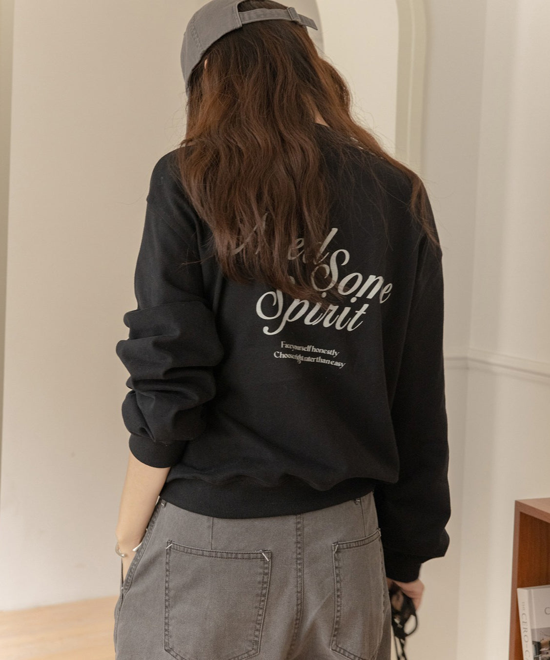 黒のスウェットを着用し、グレーのキャップと濃いグレーのデニムパンツを合わせた女性の半身ショット。背中に白いプリントロゴがある。