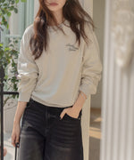 アプリコットのスウェットを着用し、黒のロングデニムスカートを合わせた女性の全身。屋外の柵のそばに立っている。