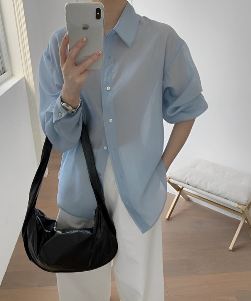ライトブルーのシアー素材の長袖シャツを白のワイドパンツに合わせ、黒のショルダーバッグをかけた女性のコーディネート。