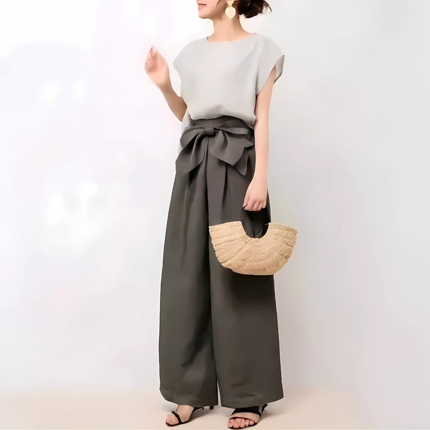 グレーのトップスとチャコールグレーのワイドパンツを着用し、ナチュラルな素材のバッグを持つ女性。リボンディテールが目を引く。