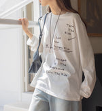 白のメッセージプリントロングTシャツとライトブルーのワイドデニムパンツを着用した女性が窓際に立った半身画像。