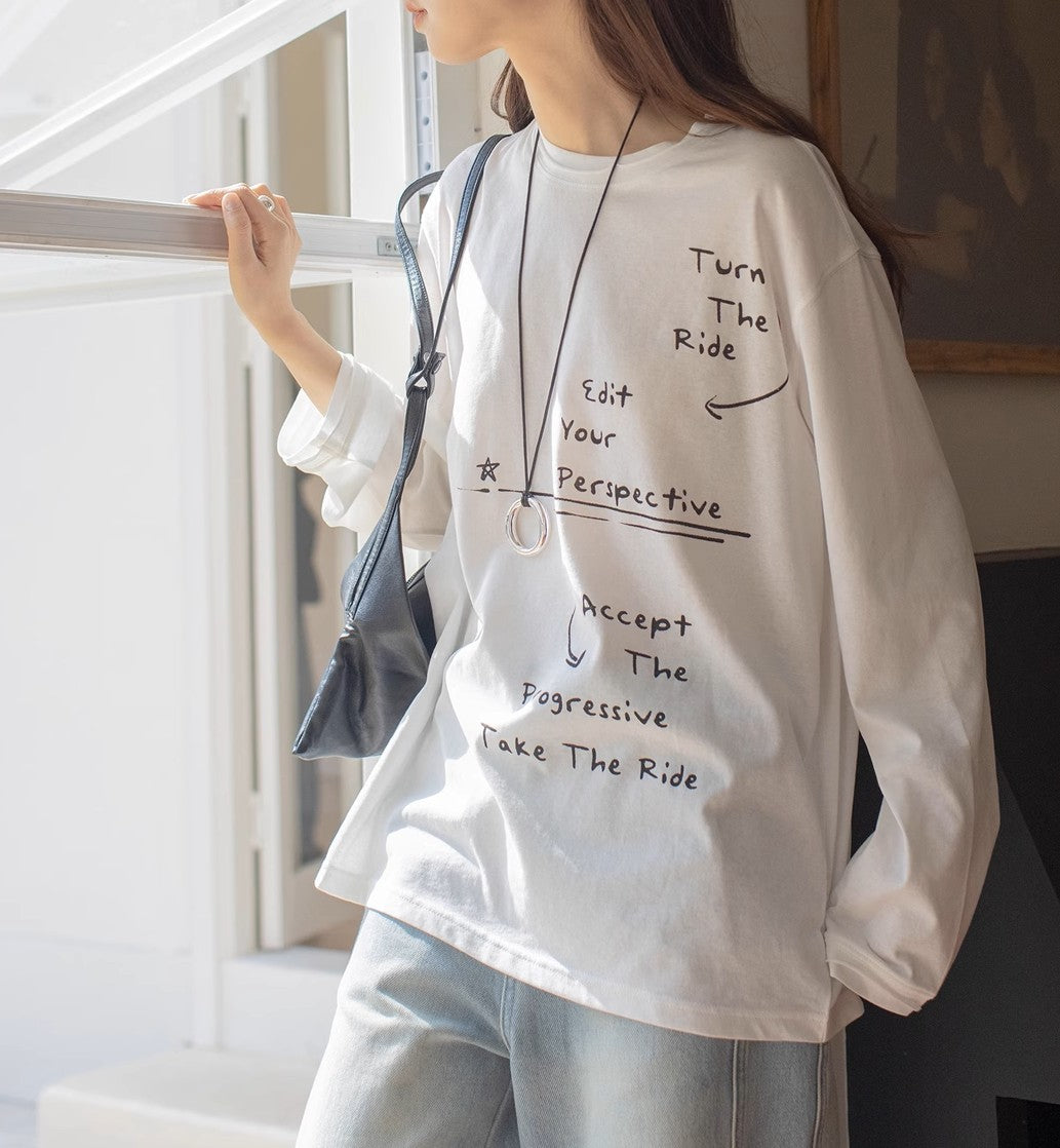 白のメッセージプリントロングTシャツとライトブルーのワイドデニムパンツを着用した女性が窓際に立った半身画像。