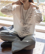 白のメッセージプリントロングTシャツとワイドパンツを着用し、窓辺に座っている女性。オーバーサイズ感が特徴的。